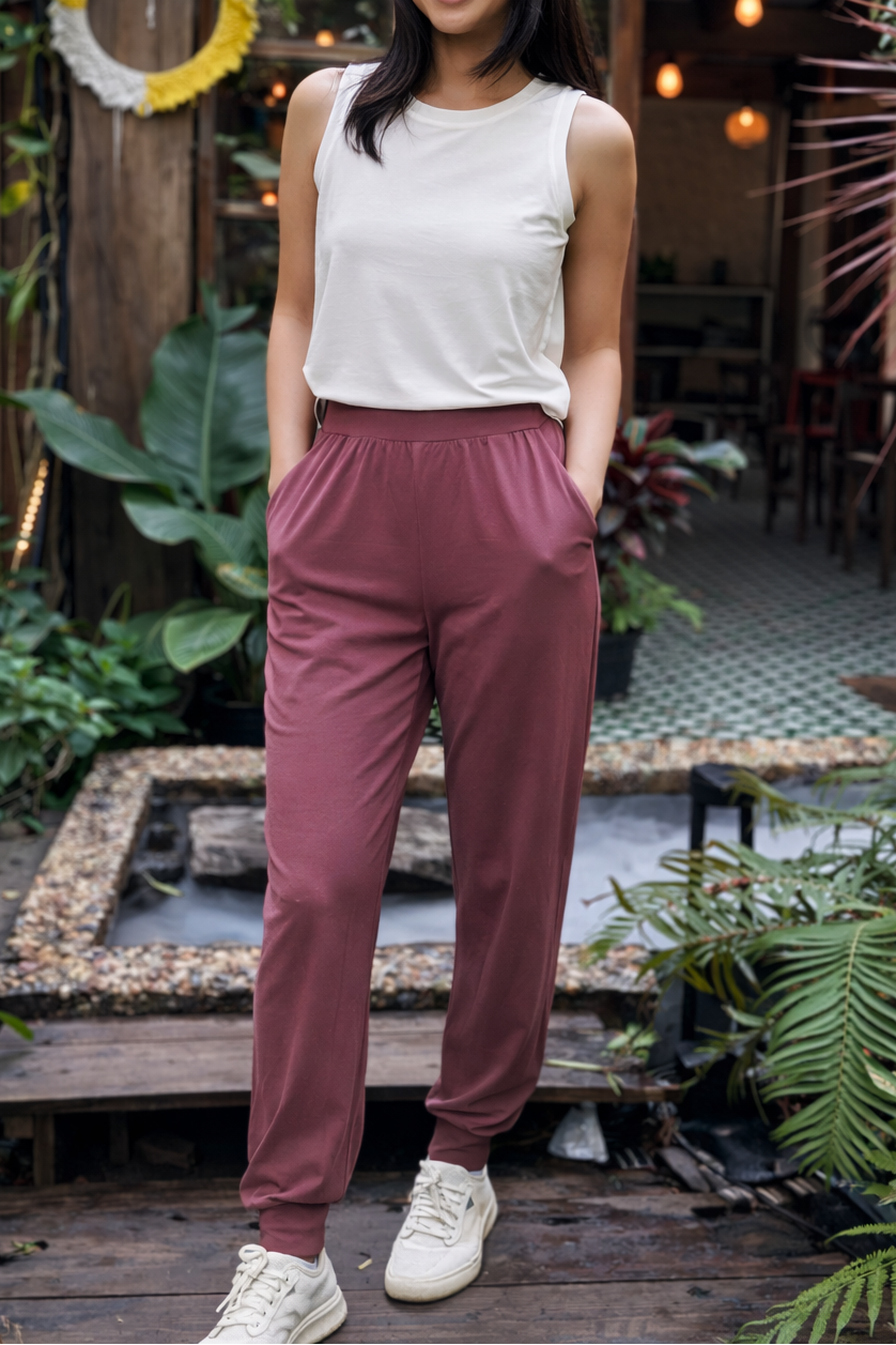 Woman in a white top and purple joggers pants standing in a garden setting with plants and wooden structures.