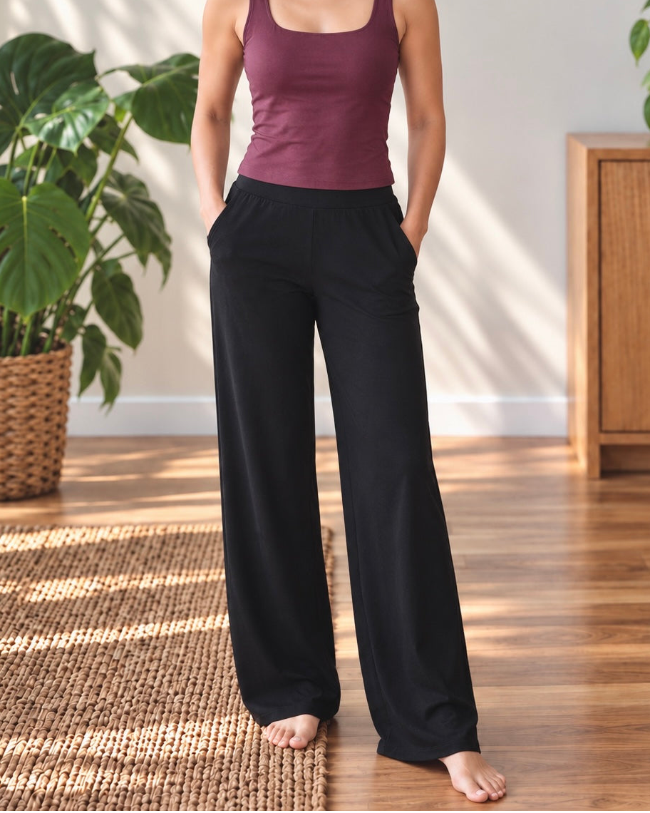A woman stands barefoot in a bright room wearing a sleeveless maroon tank top and loose black wide-leg pants, with her hands in the pockets. Large green potted plants and a wooden cabinet are visible in the background.