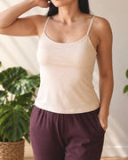 A woman wearing a camisole tank top with built-in shelf bra in sand beige and dark plum pants, standing indoors next to a green potted plant.