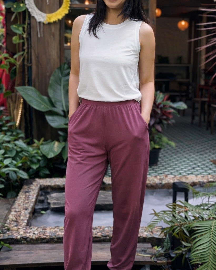 Person wearing a white sleeveless top and maroon pants standing outdoors with plants and a wooden structure in the background.