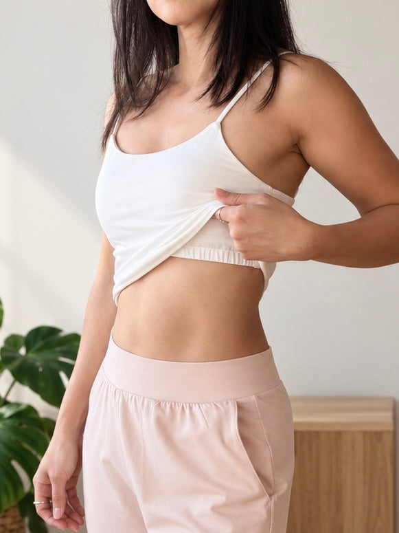 A woman wearing a white organic cotton cami tank built-in shelf bra  and light pink high-waisted shorts, lifting the top to show the fit. The photo is taken in a brightly lit indoor setting with a plant and wooden furniture in the background.